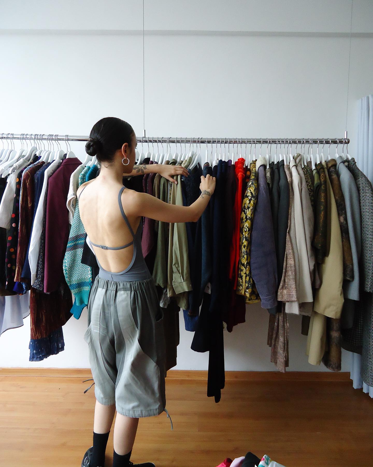 De costas, mulher mexe em arara de roupas vintage com peças coloridas. Ela usa um body com costas nuas e bermuda oversized - Metrópoles