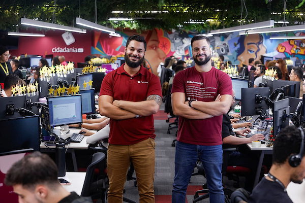 Fotografia colorida mostrando dois homens com camisa vermelha de braços cruzados dentro de sala cheia de funcionários-Metrópoles