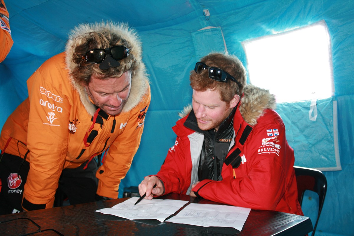 Imagem colorida mostra dois homens brancos britânicos durante excursão na Antártica. Dominic West, homem aduto, branco está usando roupa de frio laranja. ele observa o príncipe Harry, homem branco, ruivo, de roupas de frio vermelha, verificando anotações em um caderno. A foto foi tirada em 2013 - Metrópoles