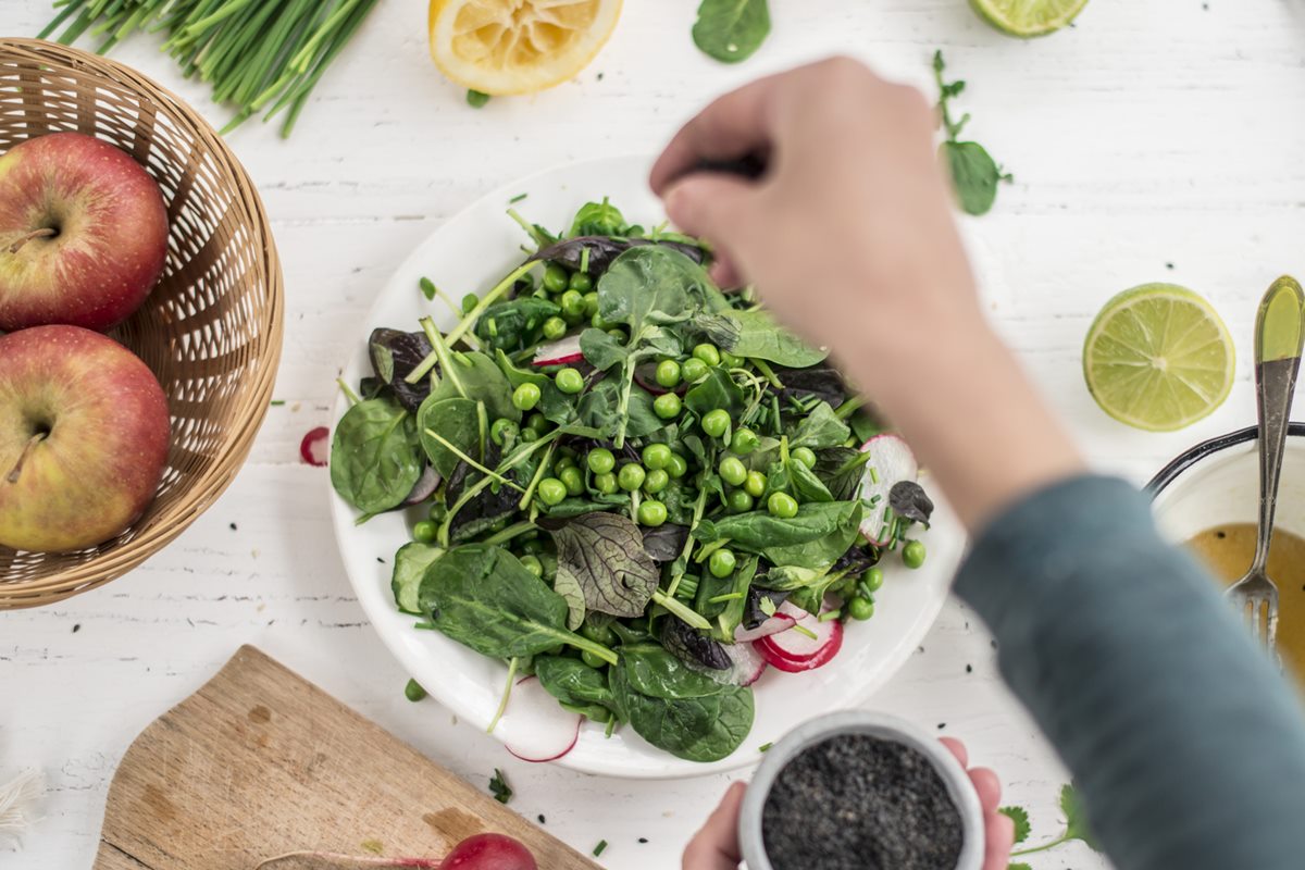 Foto colorida de prato branco com verduras e legumes, enquanto mulher coloca tempero na salada - Metrópoles