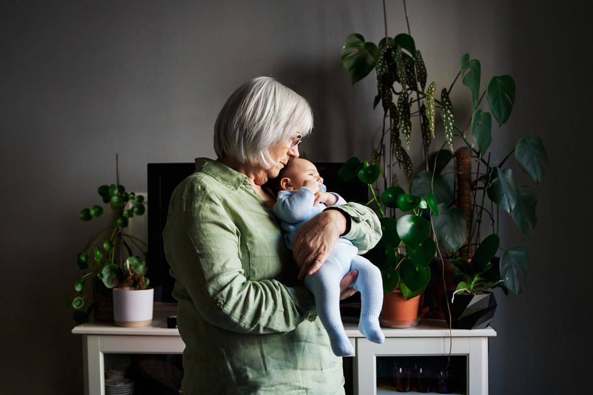 Foto colorida de mulher idosa segurando um bebê no colo. Ela está próximo de uma mesa com plantas - Metrópoles