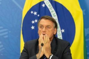 Foto colorida de Bolsonaro com mãos nas cabeça e fundo da bandeira do Brasil -- Metrópoles