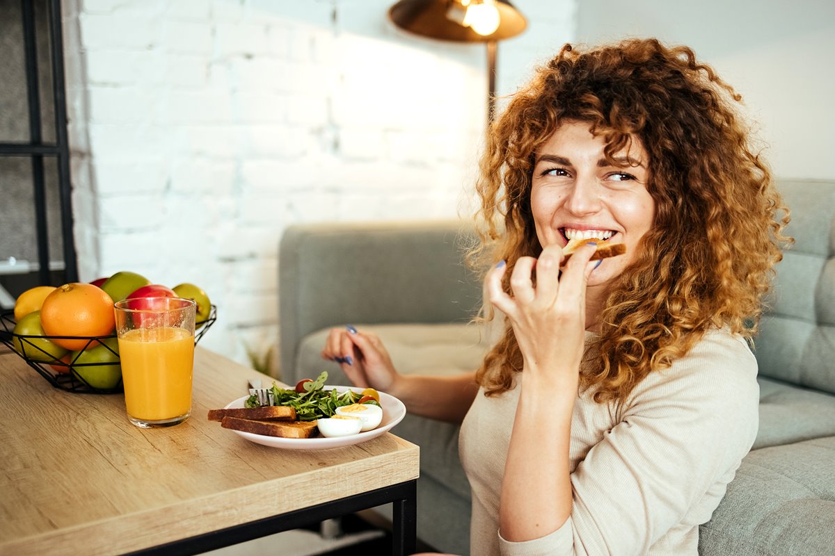 Foto de mulher comendo um pão. Ela está em frente a uma mesa com um prato, cheio de alimentos, e um copo de suco - Metrópoles