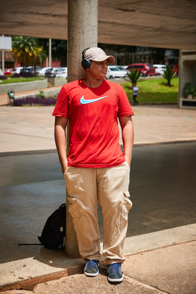 Jovem negro posa para a foto. De fone e boné como acessórios, o rapaz usa uma camiseta vermelha, calça bege e tênis azuis.