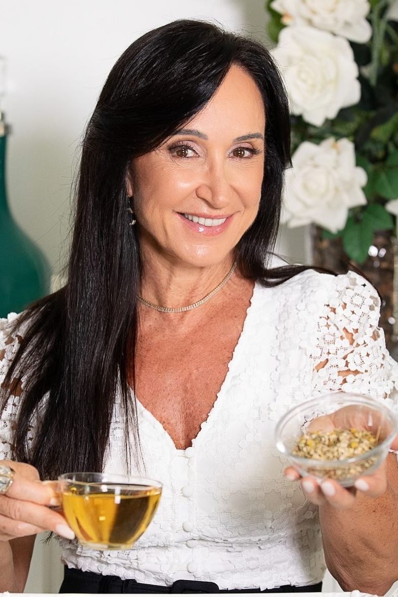 Foto de mulher com cabelo longo e preto. Ela está com uma blusa branca e segura uma xícara de chá e um pote de vidro com sementes dentro - Metrópoles