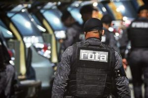 Em foto colorida policial militar (PM) de costas, na qual aparece a palavra Rota sobre o colete, com viaturas de fundo durante operação de combate à violência - Metrópoles