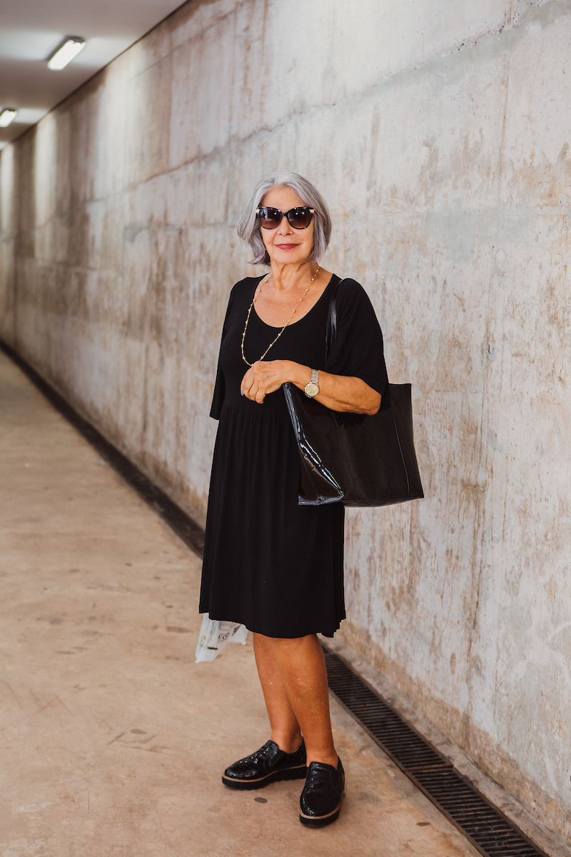 Mulher de vestido preto posa para foto. Com seu óculos escuros, ela segura sua bolsa com o ombro esquedo junto ao relógio no mesmo braço. Nos pés, a mulher calça um sapato casual loafer.