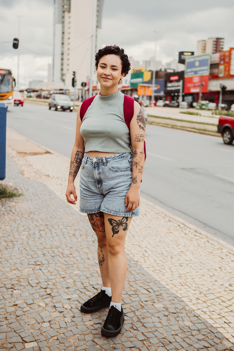 Mulher posa para foto de camiseta cropped regata e shorts. Com suas tatuagens ela esboça um leve sorriso enquanto olha para foto.