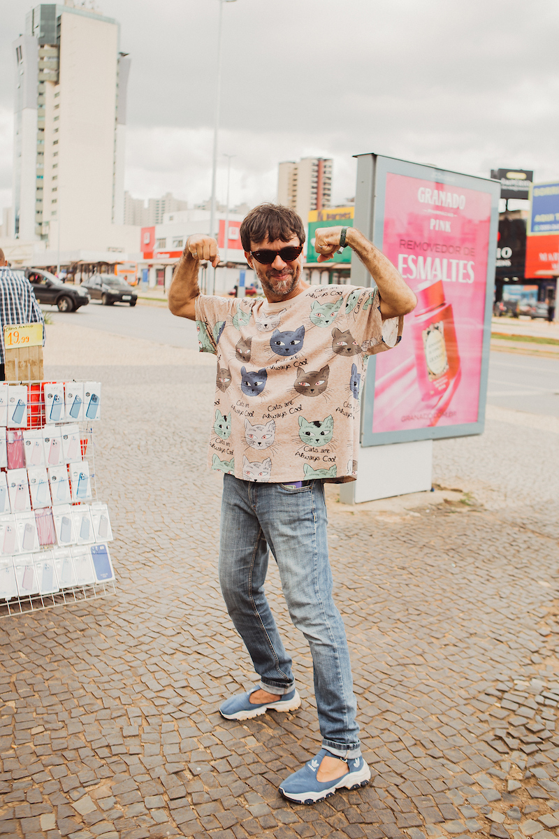 Homem faz pose imponente para foto. De óculos escuro e camiseta com estampas de gatos, ele posa em frente ao comércio local.