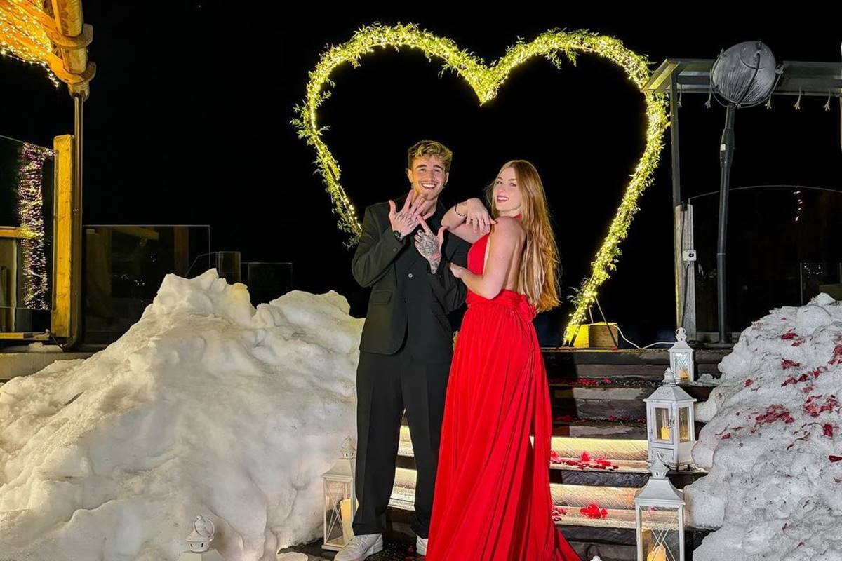 Alex Mapeli e Flávia Charallo vão se casar. Os dois estão em frente a coração luminoso, ela usa vestido vermelho e ele, terno preto - Metrópoles