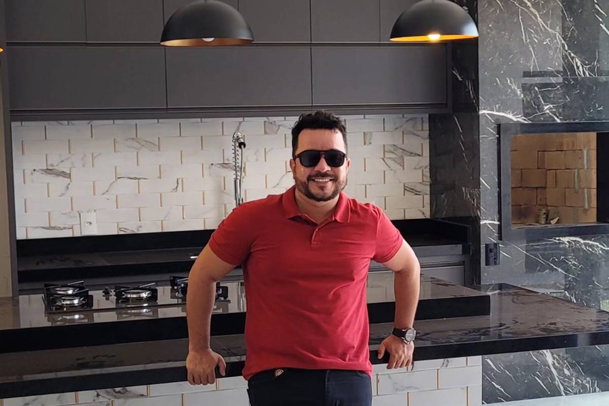 foto colorida de homem de camiseta vermelha e oculos escuros posando em cozinha - metrópoles