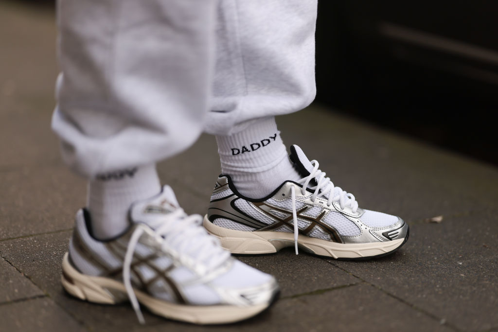 Calçado em branco e dourado aparece desamarrado. A meia aparente com a palavra Daddy estampada combina com a calça moletom também na cor branca.