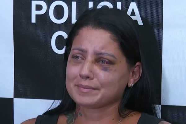 captura de tela mostra mulher com hematomas no rosto e chorando. Ao fundo, ve-se a parede da polícia civil 