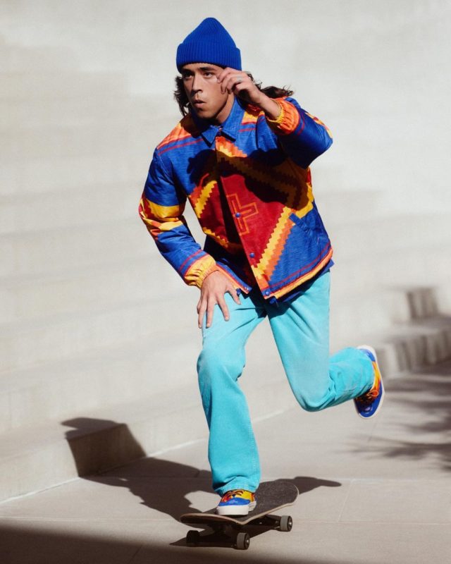 Homem é fotografado andando de skate. Na imagem, ele aparece vestindo uma camisa nas cores azul, amarelo, vemelho e laranja, combinando com seu tênis no mesmo padrão de cores, além da touca e calça azul.