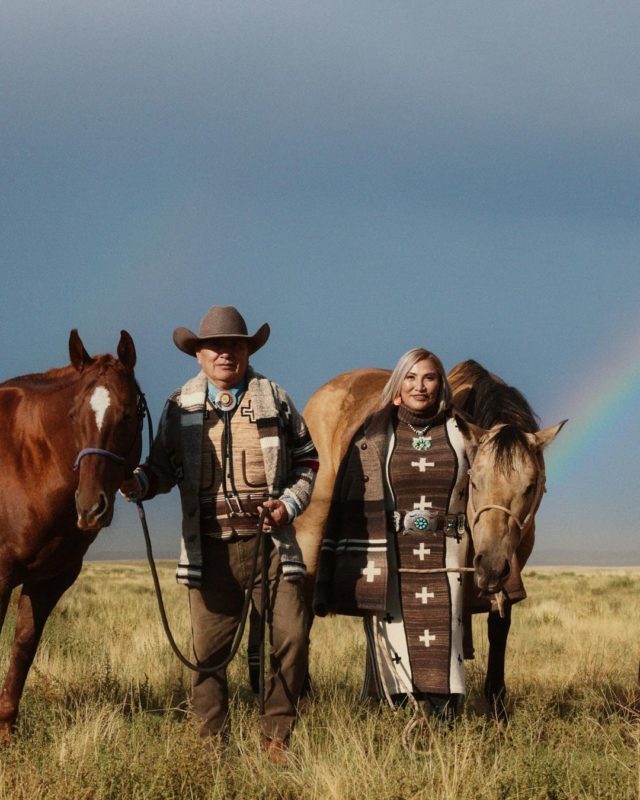 Casal fotografado com dois cavalos. Homem e mulher estão vestidos em tons terrosos com o deserto do Arizona ao fundo, acompanhado de um arco-íris no canto inferior direito da imagem.