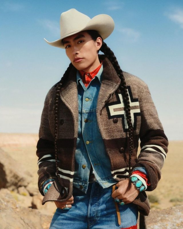 Modelo posa com casaco em tom terroso sobrepondo conjunto em jeans azul. Com seu chapéu e joias ele olha para a imagem que mostra um deserto ao fundo.