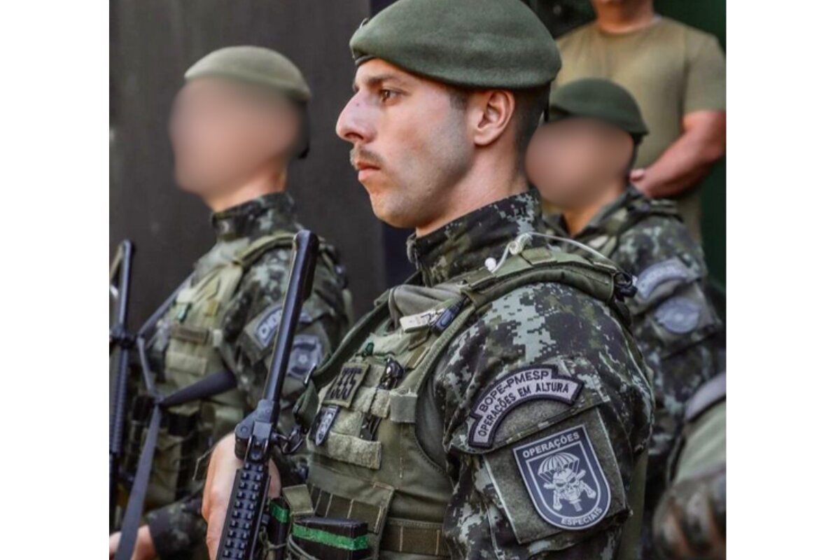 Foto colorida de PM usando farda camuflada, verde, usando boina também verde, de lado, com braçal do Grupo de Operações Especiais da PM paulista, com fuzil a tira colo - Metrópoles
