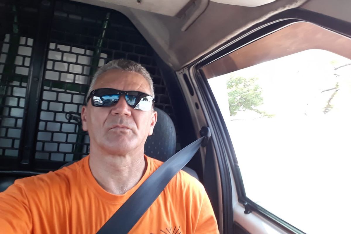 Homem branco com cabelos grisalhos curtos veste óculos de sol. Ele está com cinto de segurança, dentro de veículo de entrega.