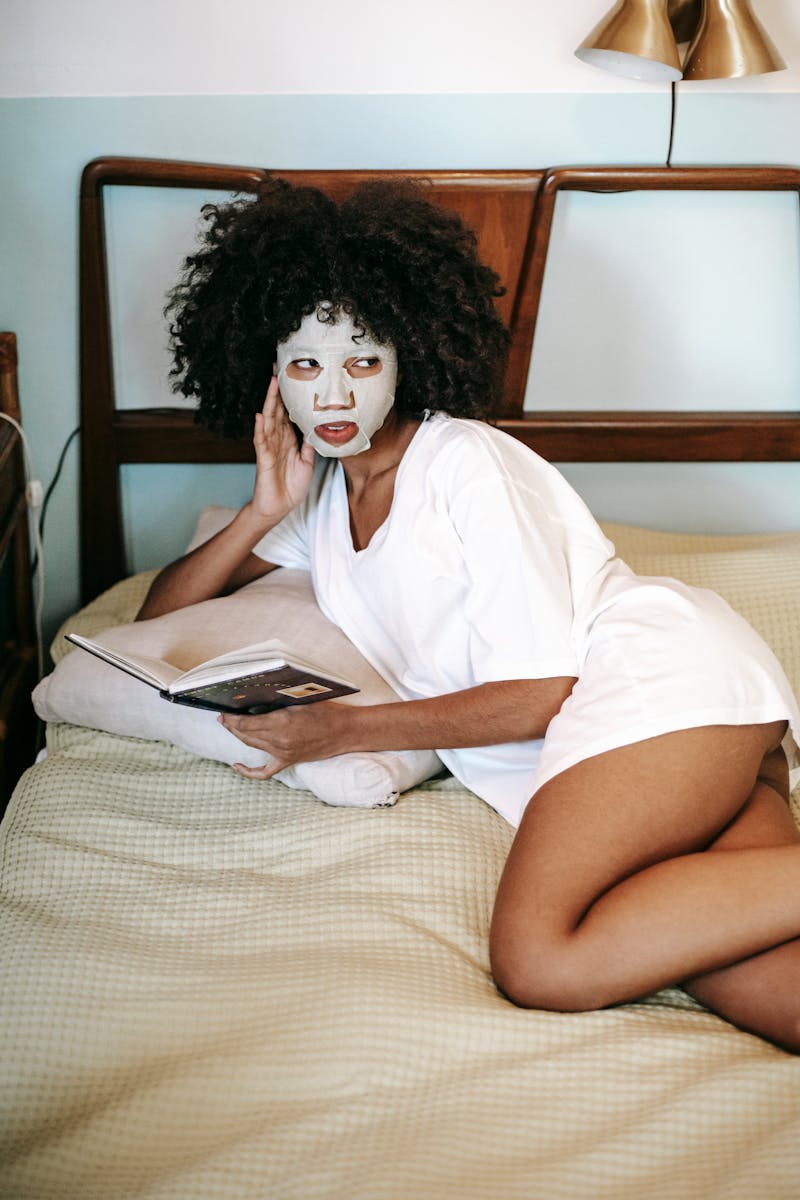 Jovem mulher com máscara branca de hidratação no rosto segurando livro deitada em cama - Metrópoles