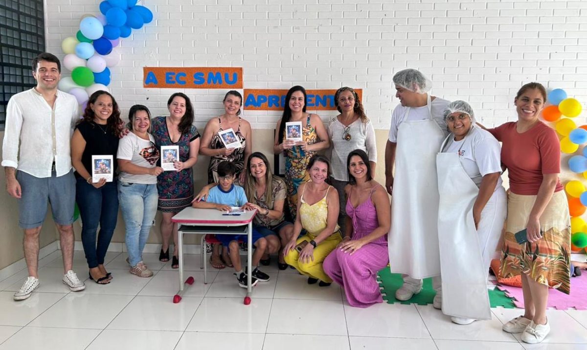 Foto colorida de adultos segurando livro em mãos ao redor de uma criança sentada de uniforme escolar