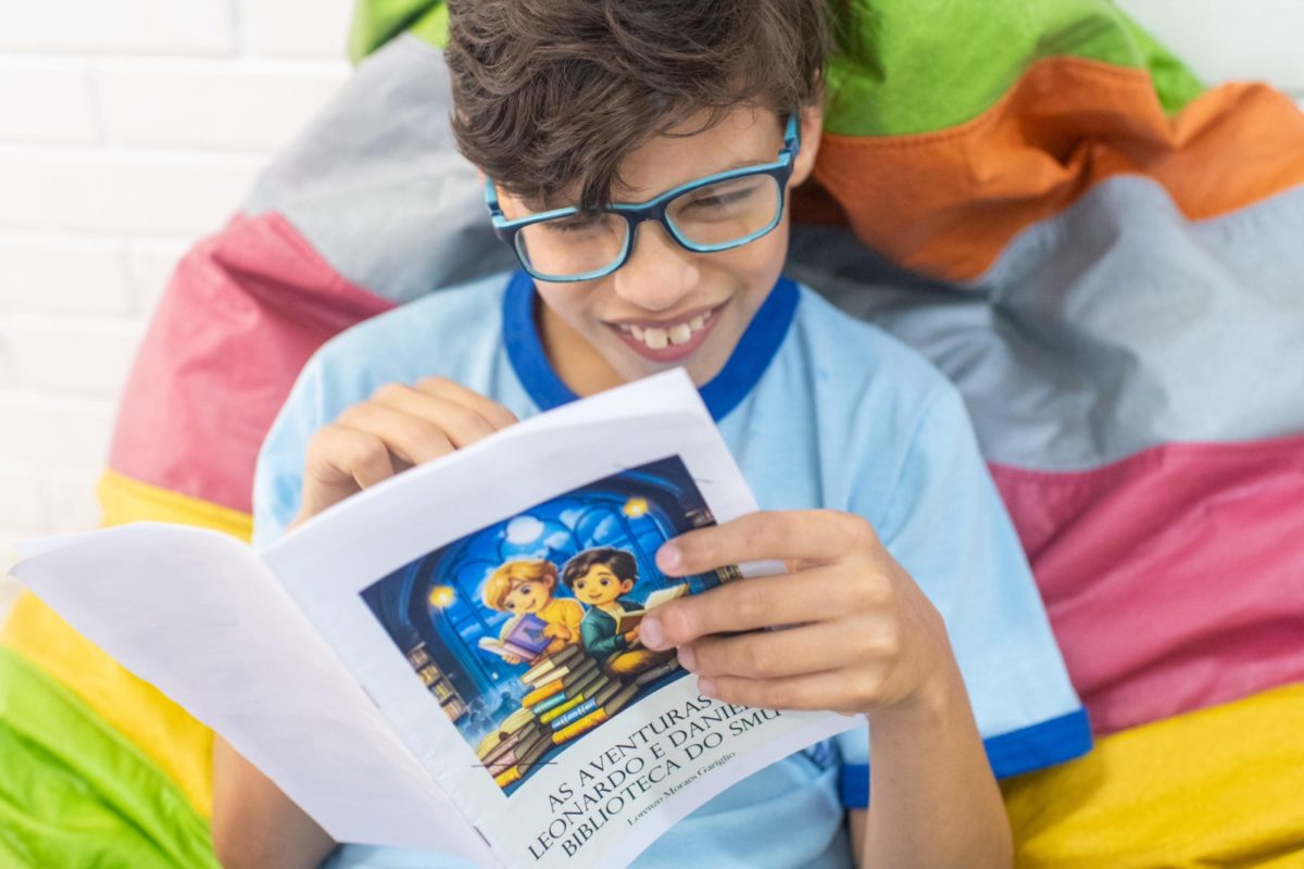Foto colorida de criança de óculos de grau, uniforme escolar e cabelos castanhos, segurando um livro com capa colorida