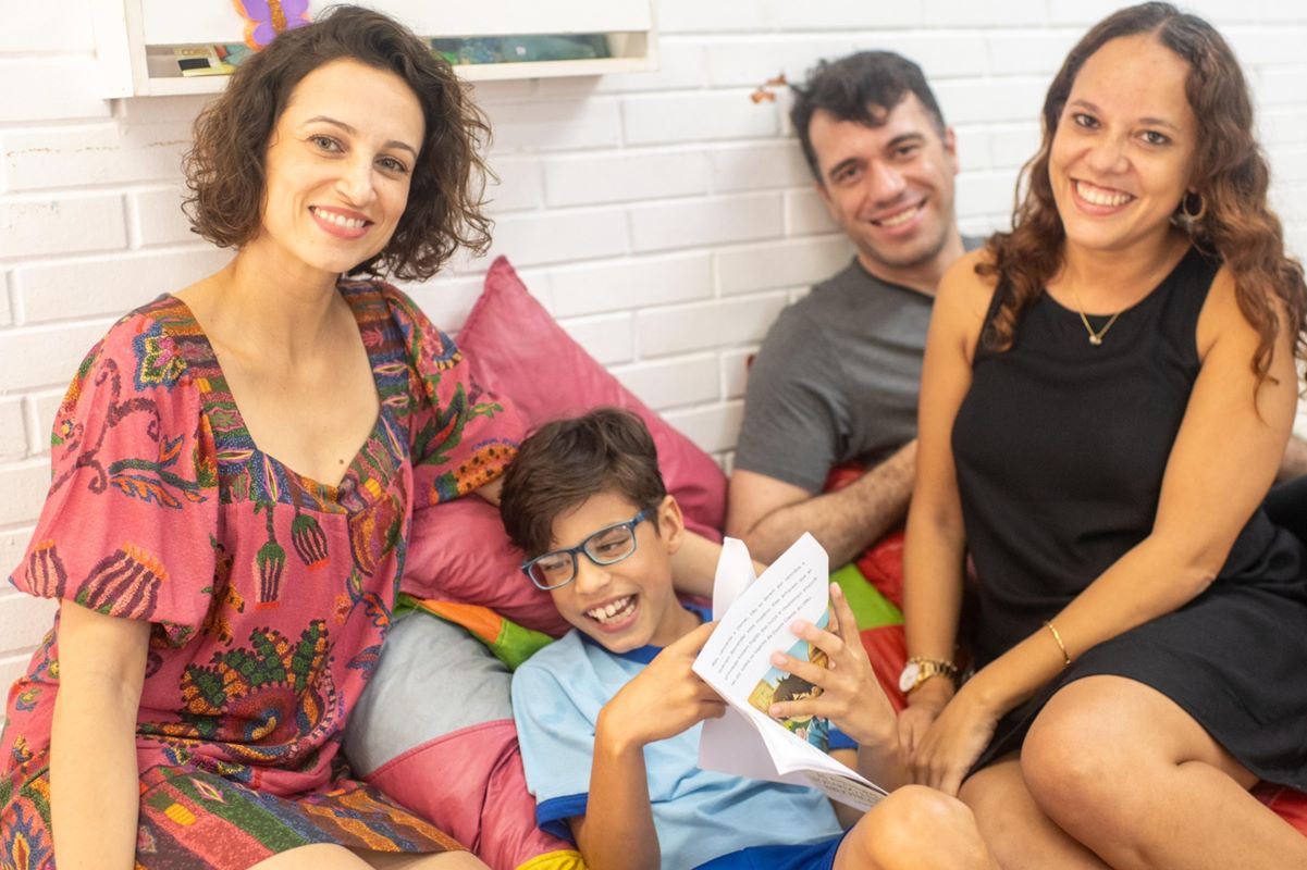 Foto colorida de uma mulher de cabelos curtos cacheados, criança de óculos de grau, homem de cabelos pretos curtos e mulher de cabelos cacheados longos, todos sentados em pufes