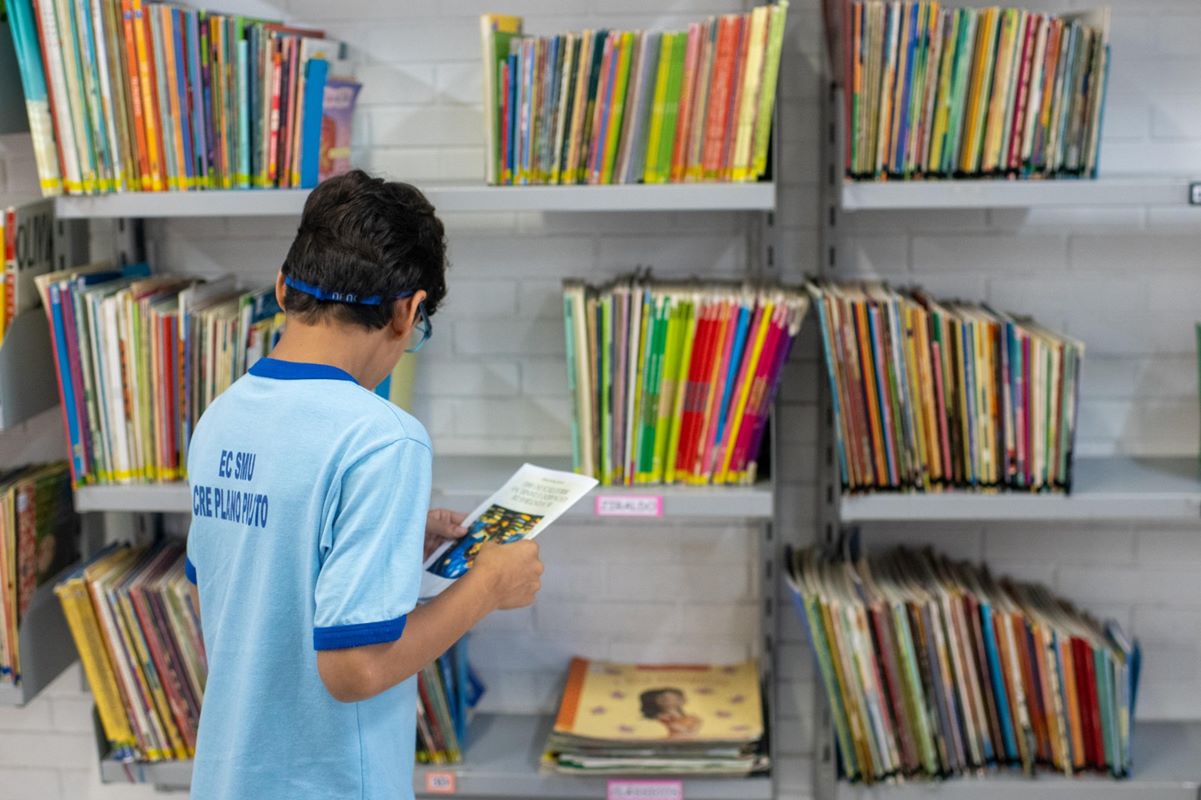 Foto colorida de uma criança de costas, uniforme de escola e cabelos curtos castanhos, de frente para uma prateleira de livros