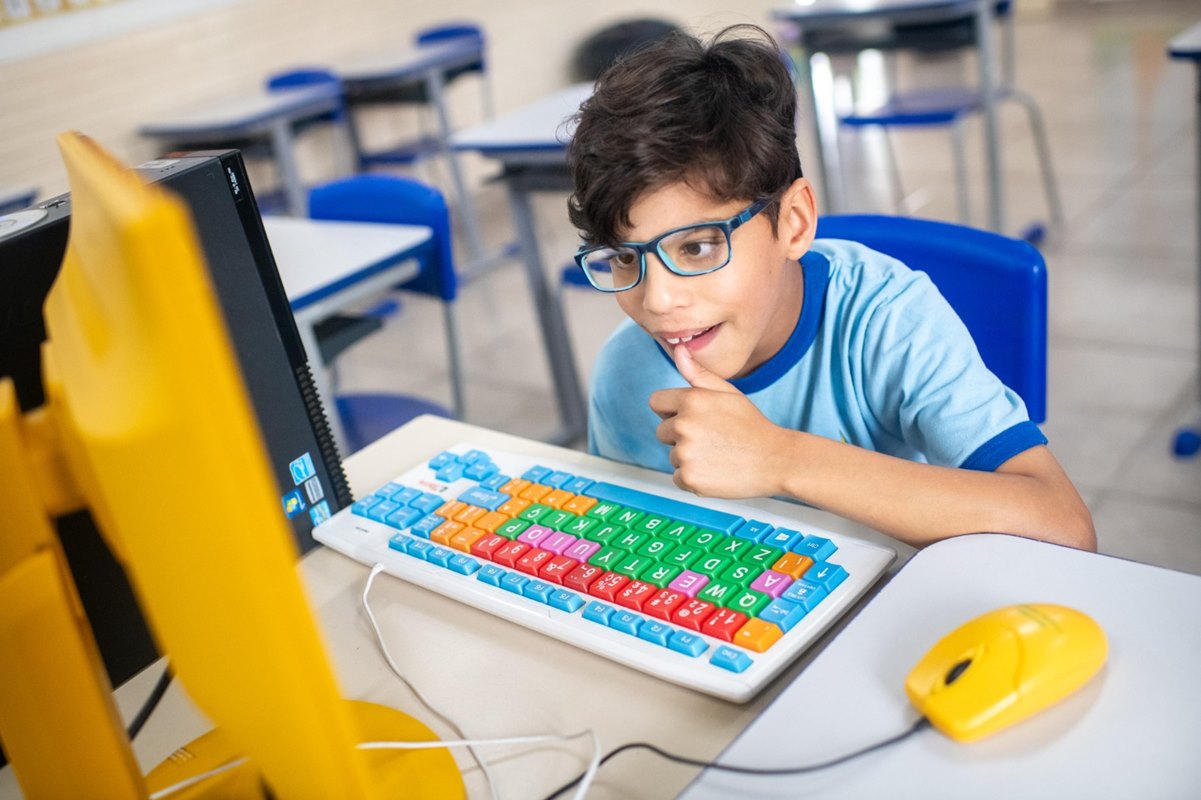 Foto colorida de criança de óculos de grau, cabelos castanhos e uniforme escolar usando um computador de teclado colorido