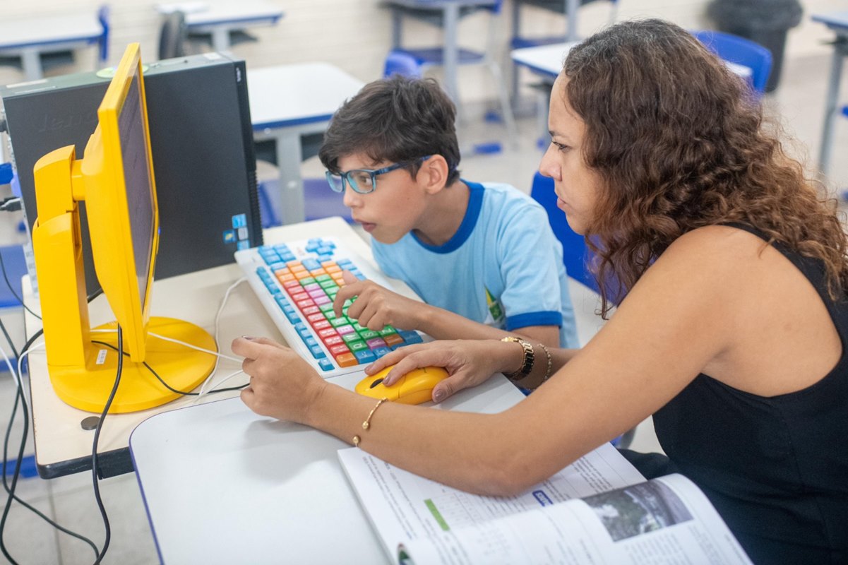 Foto colorida de criança usando um computador acompanhado de uma mulher de cabelos cacheados castanhos