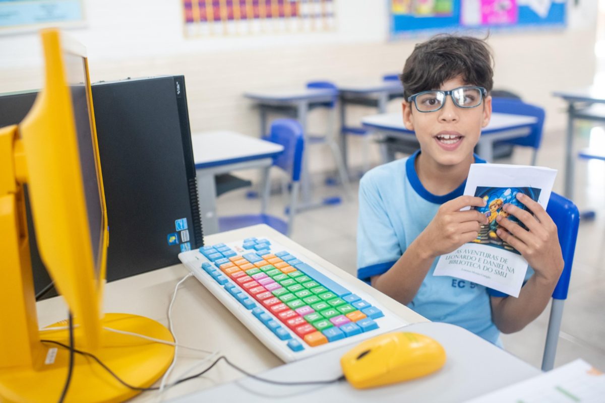 Foto colorida de criança de óculos de grau, uniforme escolar e cabelos castanhos, segurando um livro com capa colorida em frente a um computador de teclado colorido