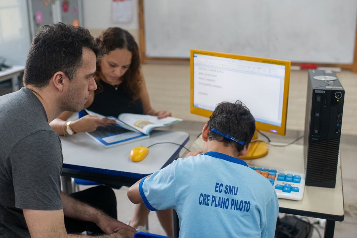 Foto colorida de criança usando um computador acompanhado de um homem de cabelos curtos pretos e uma mulher de cabelos cacheados castanhos