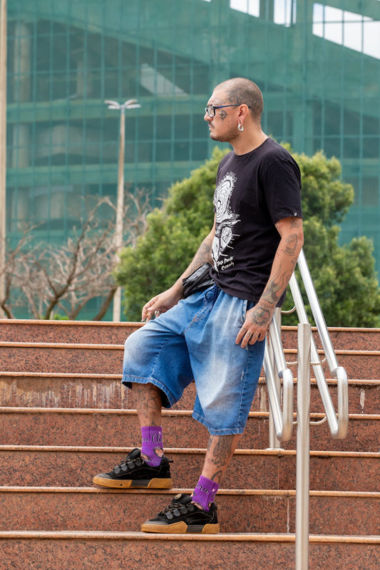 Homem posa para foto. Parado em um escada em espaço aberto, ele veste uma camisa preta com estampa branca, combinada com sua pochete na perna direita. Na parte inferior, ele usa um short jeans azul. Seu calçado preto com sola marrom, vem acompanhado de uma meia roxa com estampas em laranja.