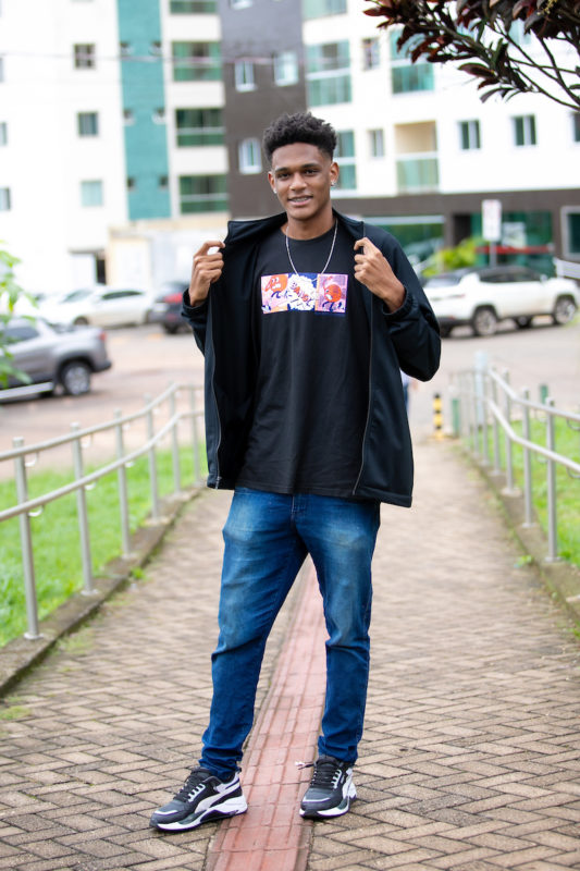 Rapaz posa para a foto. Abrindo seu casaco com zíper, ele sorri ao mostrar a estampa nas cores laranja, azul, branco e rosa em sua camisa preta. Na parte inferior, ele veste uma calça jeans, combinando com seu tênis esportivo nas cores preto e branco.