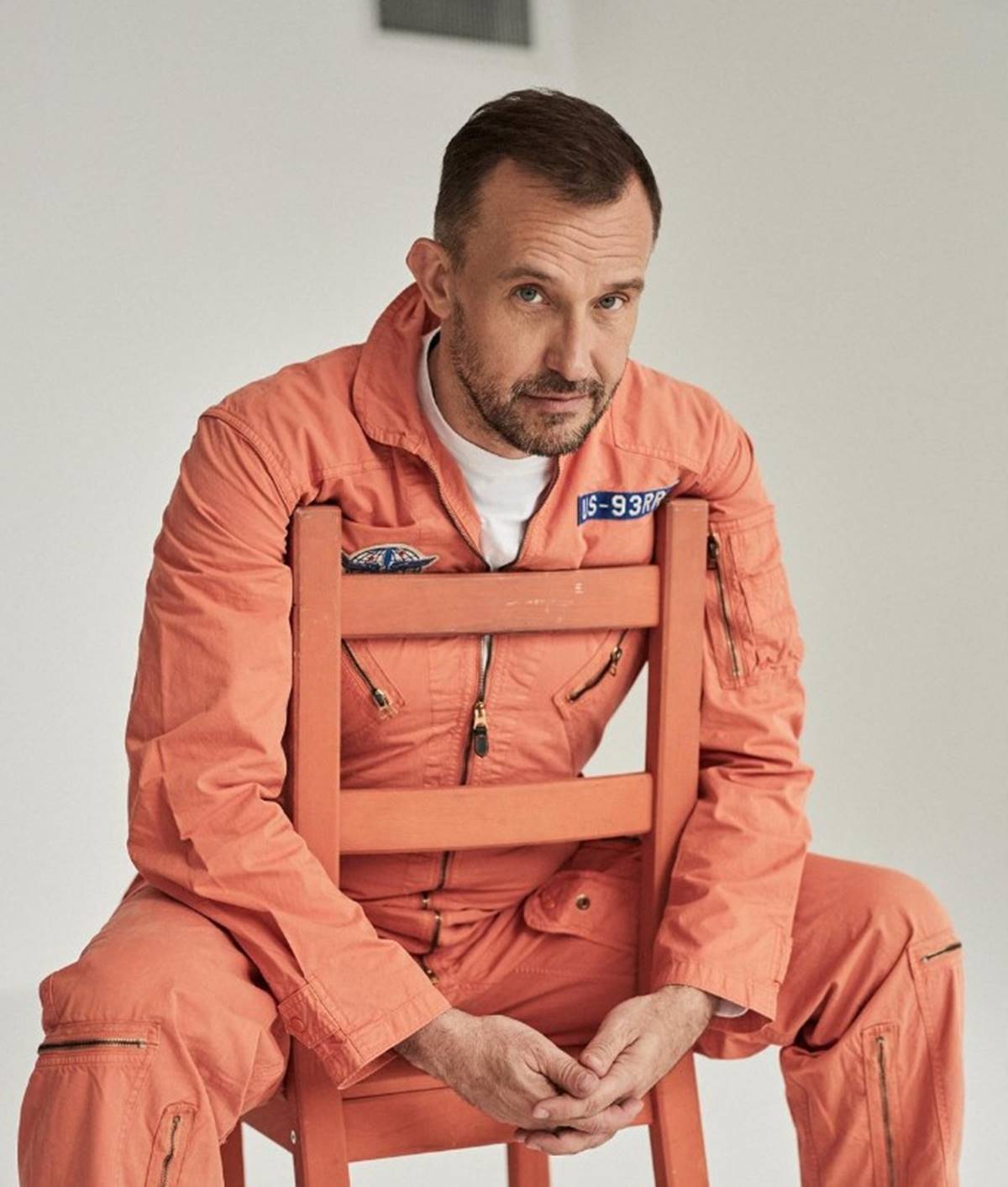 Foto colorida de um homem branco e loiro com um macacão laranja sentado em uma cadeira ao contrário em um fundo branco - Metrópoles