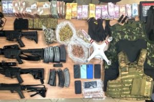Imagem colorida de objetos, armas, dinheiro e munições espalhadas em uma mesa. Metrópoles