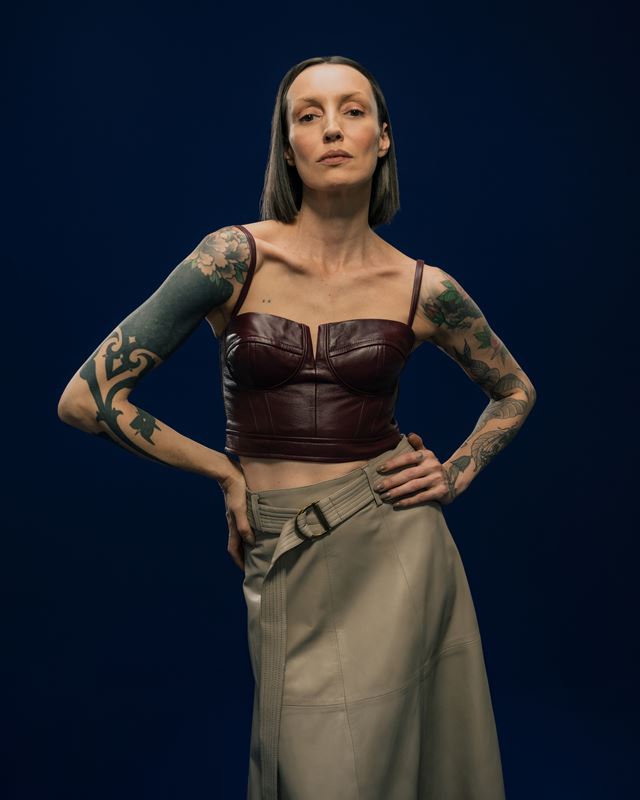Modelo tatuada posa com fundo azul. Ela usa um top marrom e saia acinzentada - Metrópoles