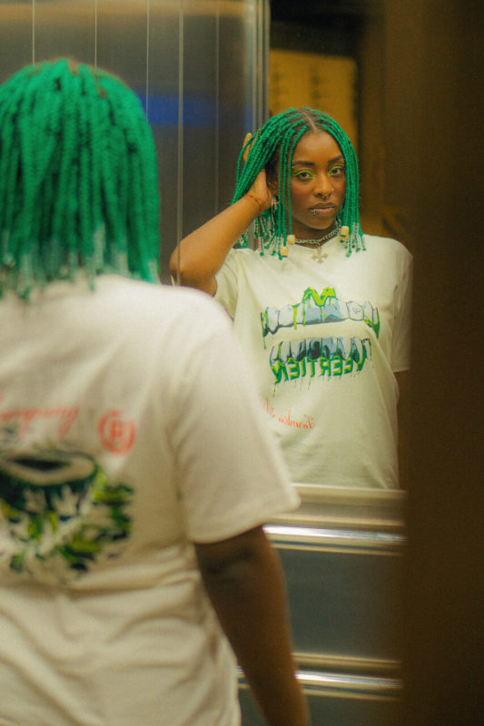 Moça posa para foto. De frente para o espelho, ela direciona seu olhar para câmera enquanto mexe em seu cabelo verde exibindo a camiseta.