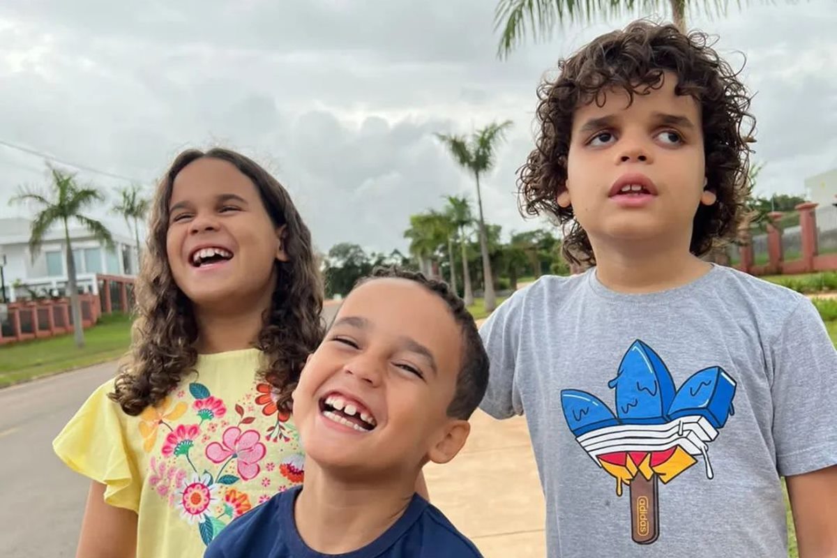 Foto colorida de três crianças posando para foto. Elas estão sorridentes - Metrópoles